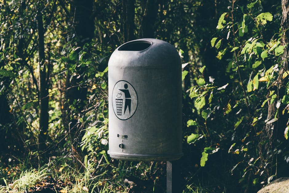 A metal trash can stands amidst vibrant, lush greenery in a peaceful forest.
