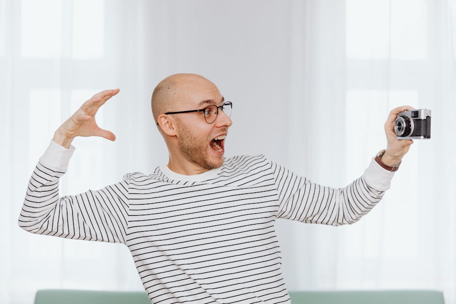 Bald man with eyeglasses playfully takes a selfie while posing indoors.