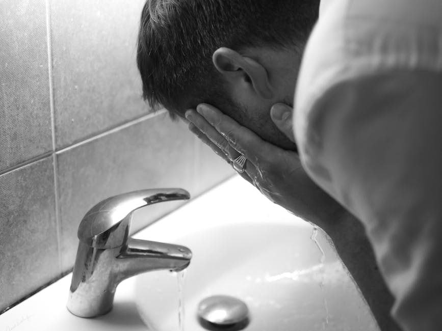 Monochrome image of a man washing his face at a sink with running water.