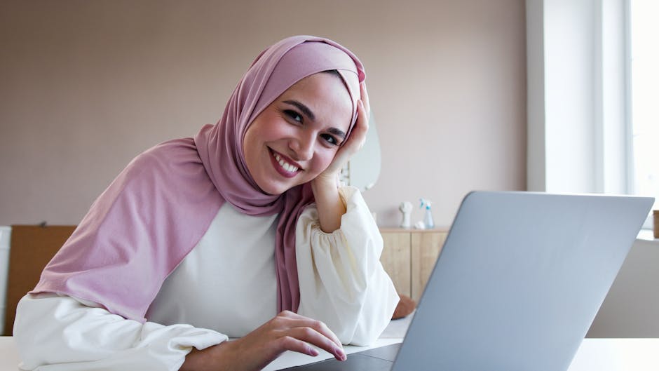 Professional woman in hijab smiling while working on a laptop in a modern office setting.