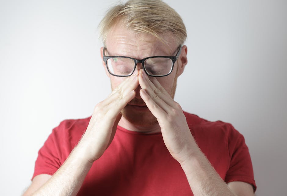 Tired man with glasses rubbing eyes, expressing stress or fatigue in casual red shirt.