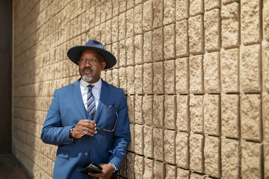 Confident businessman in blue suit and hat leaning against textured wall outdoors.