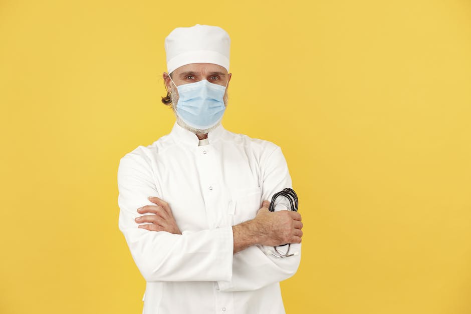 Confident male doctor with arms crossed wearing a surgical mask against a vibrant yellow background.