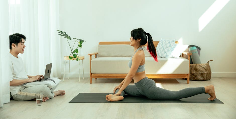 A woman practices yoga while a man uses a laptop in a bright living room.