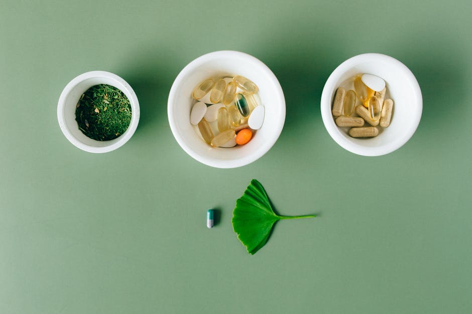 Flatlay of capsules and herbs in ceramic bowls on green surface.