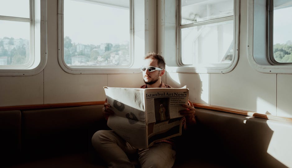 A man wearing sunglasses reads a newspaper on a boat in Istanbul, Türkiye during daylight.