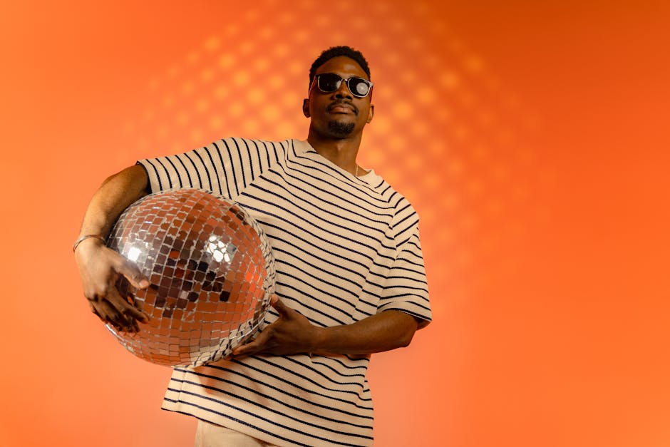 A fashionable man wearing sunglasses holds a disco ball against an orange studio backdrop.